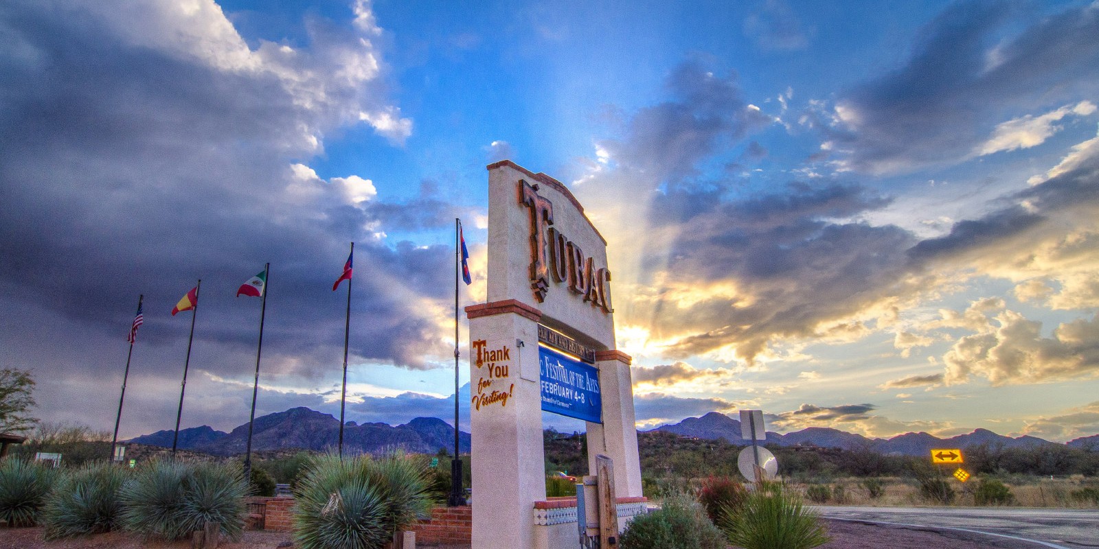 Vibrant rays of the setting sun emerge from behind the clouds. In the foreground, a sign welcoming people to the village of Tubac, Arizona.